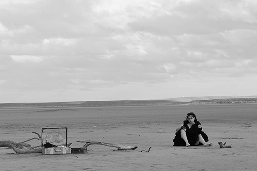 In a vast, flat sandy landscape, a woman in black dress sits with open suitcase