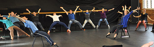 In a dance studio, a group of 1-–20 seated male and female participants lean far back in their chairs, with arms and legs fully outstreched, pointed toes and mouths wide open.