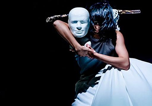 A female performer clasps hands around a white-masked performer