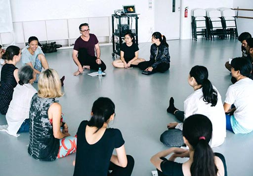 Facing the workshop leader, a circle of dancers sit cross-legged on floor.