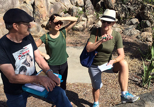 A man and two women stand in front of a rocky outcrop in a native parkland setting. The sun is harsh and one woman shields her eyes from the glare with her hands. They all wear shorts and t-shirts and other woman is faces them talking.