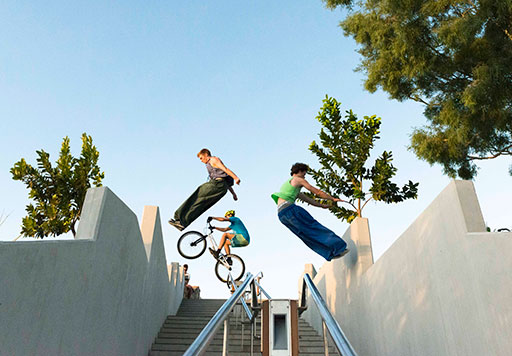 Three young men perform Parkour on a flight of steps. Two jump from the steps' concrete wall railings while one balances his bicycle on a steel hand rail.