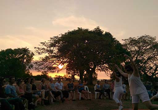 An audience sits on chairs in a circle around dancers dressed in white. Behind them, a large park tree is silhouetted as the sun sets over the sea behind them.