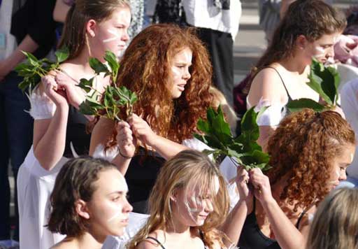 A group of young dancers perform in the middle of a large crowd. They hold branches to their shoulders.