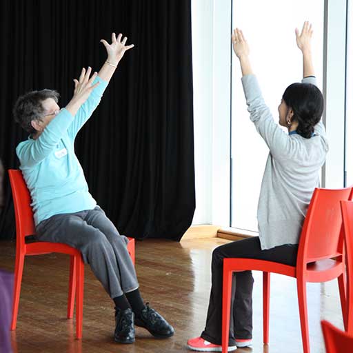Two women, one middle aged, the other in her twenties, sit on red chairs facing each other with arms streched above their heads and fingers spread wide.