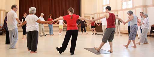 In a Dance for Parkinson's class, a group of about 20 participants of various ages stand in a circle holding each other's outstretched hands. 