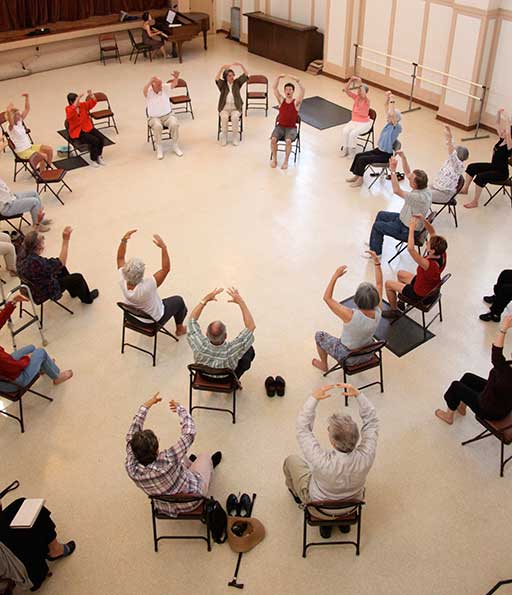 Arranged in two large concentric circles, seated with their shoes off and arms raised in a curve above their heads, 15–20 participants follow the instructors movement.