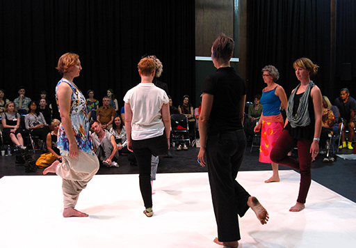 A dancer stands one-legged on the dance space. Other dancers stand in front of her, each lifting one leg up behind them to stand on one leg. They are surrounded by a watching audience, some seated while others sit cross-legged on the floor.
