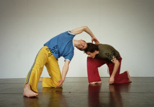Two men dancing in a studio, both bend and kneel with one reaching over to touch the others back.