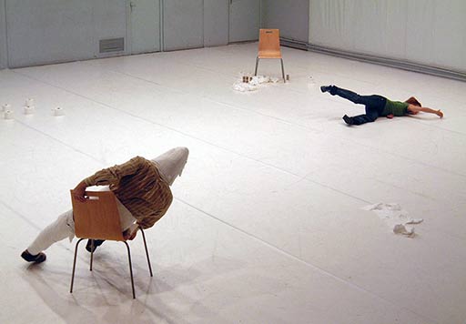 A performer leans sideways on a chair while a female dancer moves across the floor